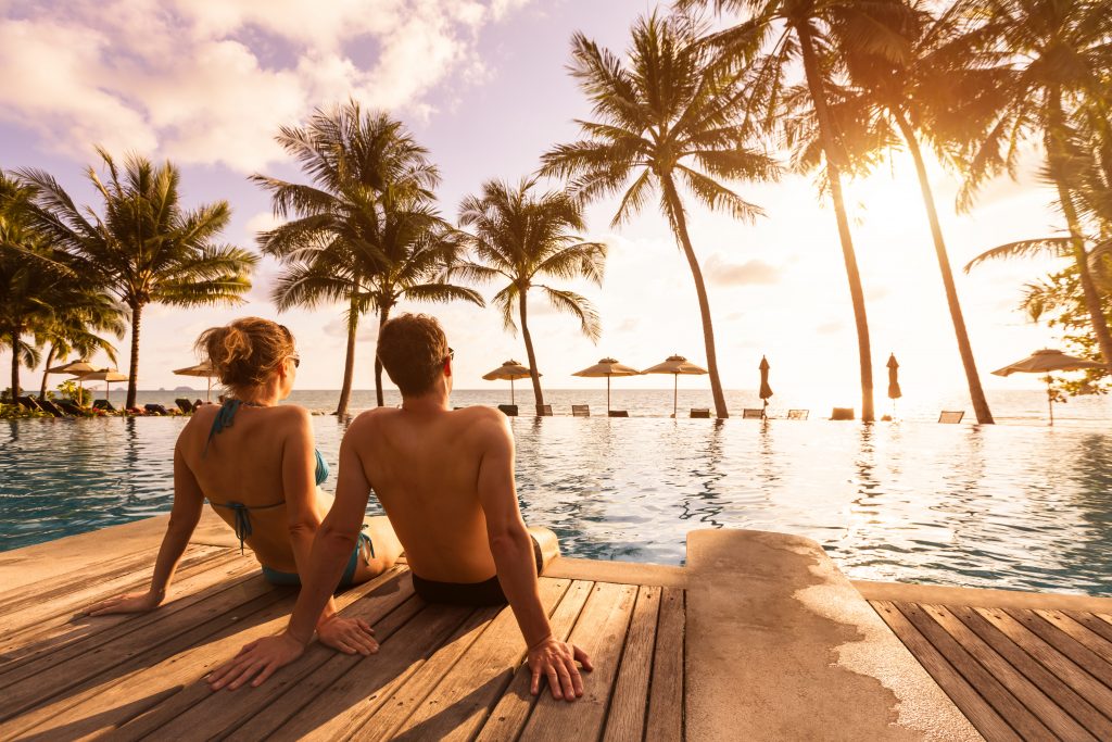Couple sitting by tropical resort pool at sunset, overlooking the ocean and palm trees.